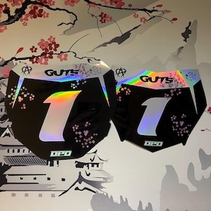May include: Two black motorcycle number plates with a holographic finish. Each plate features a large holographic number one in the centre, the text GUTS at the top, and the text DPD at the bottom. The design includes small holographic cherry blossom patterns.
