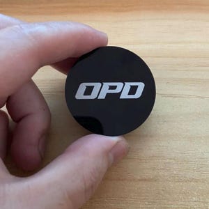 May include: A circular black disc featuring the letters OPD printed in a bold, white, sans-serif font centered on the surface. The object has a smooth, reflective finish and a clean, minimalist design, presenting a clear and legible typographic display.