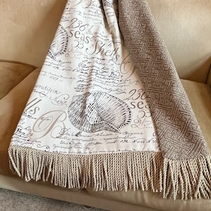 Peut inclure: Une couverture en jetée crème et marron avec une bordure en franges. Un côté présente un fond blanc avec une écriture noire et un motif de coquillage. L'autre côté est un motif géométrique marron et crème.