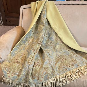 May include: A decorative throw blanket draped over a beige sofa. The blanket features a paisley pattern in shades of yellow, blue, and brown, with a solid yellow backing and cream-colored fringe along the edge. The sofa is in a room with a rug and wooden furniture.