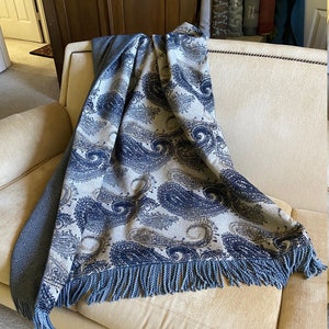 May include: A blue and beige paisley patterned throw blanket with fringe on a light beige couch.