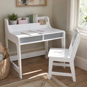 May include: A white wooden desk with two gray fabric drawers and a matching white chair. The desk features an upper shelf holding a potted green plant, a pink organizer with pens, books, and a wooden block calendar displaying the date 12 April.