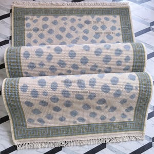 Könnte beinhalten: Beige und blau gemusterter Teppich mit rechteckiger Form und gerollten Abschnitten. Der Teppich hat ein sich wiederholendes Muster aus blauen Kreisen auf beigem Hintergrund, umrahmt von einem grünen Rand. Der Teppich hat einen Fransenrand.