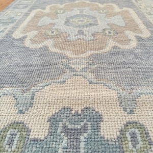 Op de afbeelding: Een rechthoekig tapijt met een gedetailleerd, symmetrisch patroon. Het tapijt heeft een centraal ontwerp in beige en bruin, omgeven door een lichtblauw veld. De rand bevat accenten van groen en blauw. Het tapijt is gemaakt van een geweven materiaal.