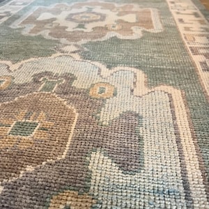 Peut inclure: Gros plan sur un tapis rectangulaire avec un motif complexe. Le tapis présente une combinaison de couleurs sourdes, notamment du vert, du beige et du marron. Le motif comprend des éléments géométriques et floraux, créant un aspect texturé.