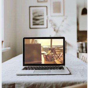 May include: A silver laptop sits open on a white tablecloth, displaying a sunny outdoor scene. The laptop is the focal point, with a keyboard and screen visible. The background includes framed artwork and a glimpse of a kitchen, suggesting a home office setup.