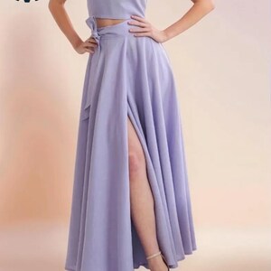 May include: A two-piece lavender outfit featuring a crop top and a high-waisted, flowing skirt with a thigh-high slit. The top has a scoop neckline. The outfit is paired with neutral-toned heels.