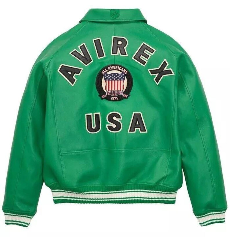 K&ouml;nnte beinhalten: Gr&uuml;ne Lederjacke mit dem Wort "AVIREX" auf dem R&uuml;cken, &uuml;ber einem runden Aufn&auml;her mit der amerikanischen Flagge und den Worten "ALL AMERICANS ESTABLISHED 1975". Das Wort "USA" steht darunter. Die Jacke hat einen wei&szlig;-gr&uuml;nen gestreiften Saum.