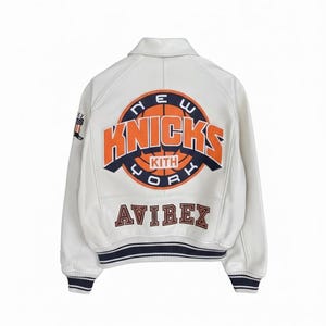 K&ouml;nnte beinhalten: Cremefarbene Lederjacke mit dem Logo der New York Knicks in Orange und Marineblau. Das Wort "Knicks" und "New York" sind sichtbar. Das Avirex-Logo ist ebenfalls auf der Jacke zu sehen.