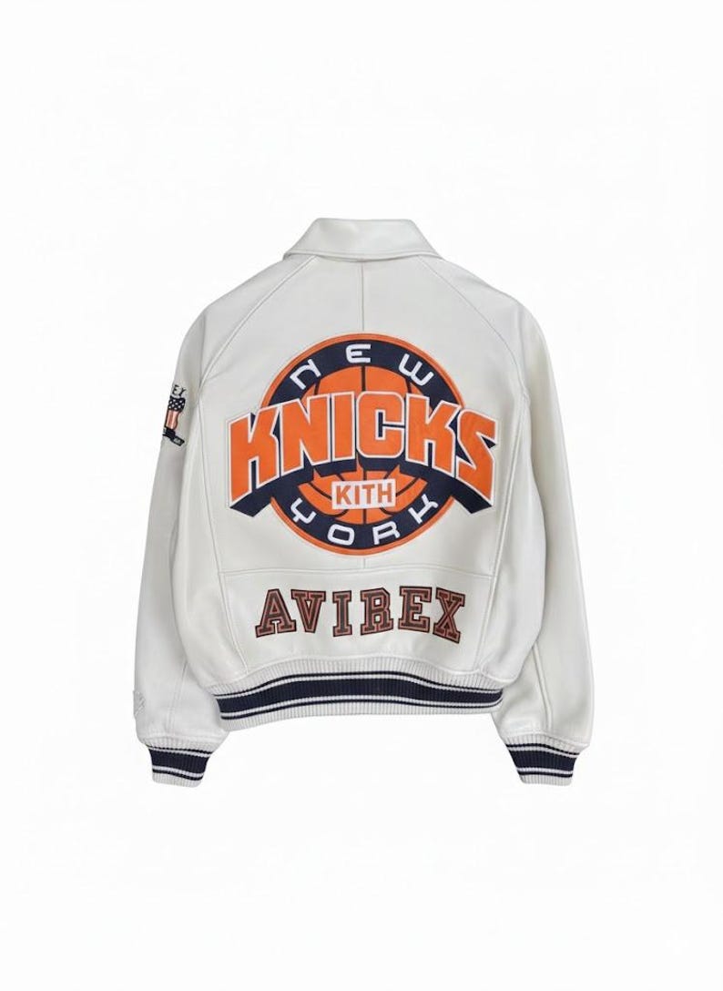 K&ouml;nnte beinhalten: Cremefarbene Lederjacke mit dem Logo der New York Knicks in Orange und Marineblau. Das Wort "Knicks" und "New York" sind sichtbar. Das Avirex-Logo ist ebenfalls auf der Jacke zu sehen.