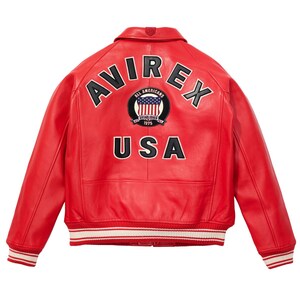 K&ouml;nnte beinhalten: Rote Lederjacke mit dem Wort "AVIREX" &uuml;ber den Schultern und "USA" darunter. Ein kreisf&ouml;rmiger Aufn&auml;her mit der amerikanischen Flagge und den Worten "ALL AMERICANS ESTABLISHED 1975" ist zentriert. Die Jacke hat wei&szlig;-rot gestreifte B&uuml;ndchen und Saum.