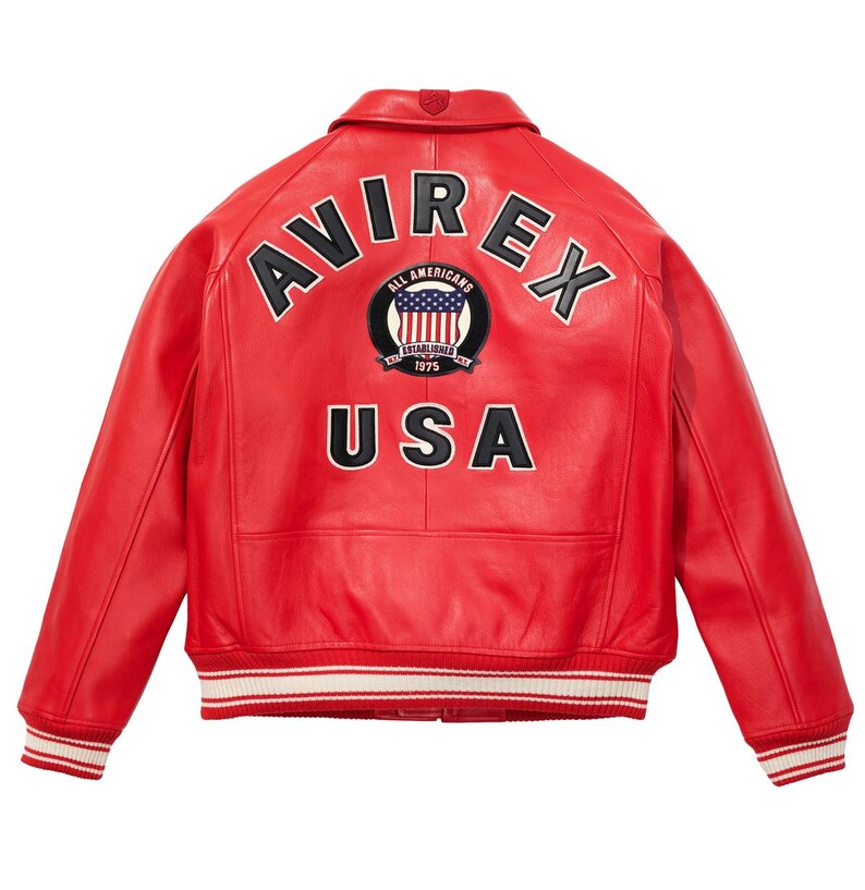 K&ouml;nnte beinhalten: Rote Lederjacke mit dem Wort "AVIREX" &uuml;ber den Schultern und "USA" darunter. Ein kreisf&ouml;rmiger Aufn&auml;her mit der amerikanischen Flagge und den Worten "ALL AMERICANS ESTABLISHED 1975" ist zentriert. Die Jacke hat wei&szlig;-rot gestreifte B&uuml;ndchen und Saum.