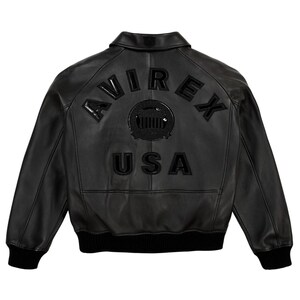 K&ouml;nnte beinhalten: Schwarze Lederjacke mit dem Wort "AVIREX" auf dem R&uuml;cken, &uuml;ber einem kreisf&ouml;rmigen Emblem und dem Wort "USA". Die Jacke hat einen gerippten Saum und B&uuml;ndchen sowie einen klassischen Kragen.