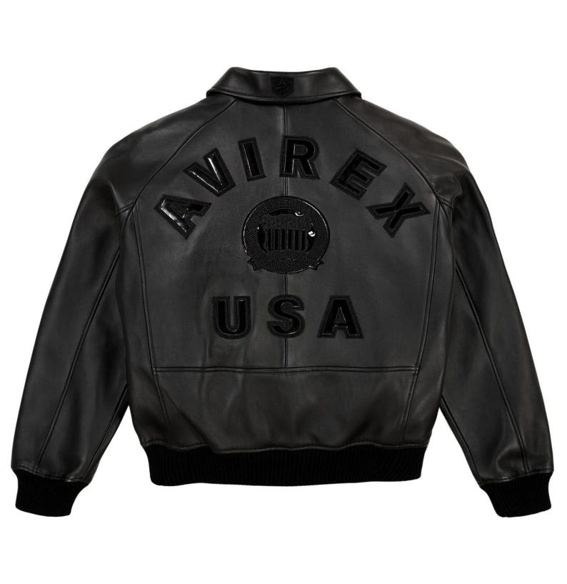K&ouml;nnte beinhalten: Schwarze Lederjacke mit dem Wort "AVIREX" auf dem R&uuml;cken, &uuml;ber einem kreisf&ouml;rmigen Emblem und dem Wort "USA". Die Jacke hat einen gerippten Saum und B&uuml;ndchen sowie einen klassischen Kragen.