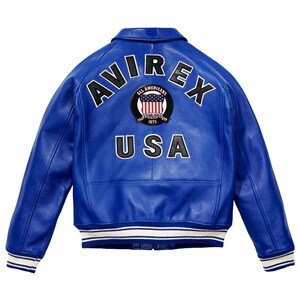 K&ouml;nnte beinhalten: Eine leuchtend blaue Lederjacke mit dem Wort "AVIREX" in wei&szlig;er Schrift auf dem R&uuml;cken. Ein kreisf&ouml;rmiger Aufn&auml;her zeigt die amerikanische Flagge und die Worte "ALL AMERICANS ESTABLISHED 1975". Das Wort "USA" ist darunter gedruckt.