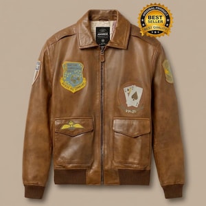 May include: A brown leather bomber jacket with a zip-up front, two front pockets, and a ribbed hem and cuffs. The jacket features embroidered patches, including a shield with text and a playing card design. The jacket has the text "AVIREX" on the label.