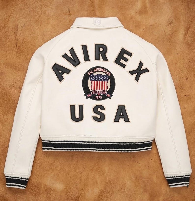 May include: Cream-colored bomber jacket with black and white striped trim. The back features the word "AVIREX" above a circular emblem with the American flag and the text "USA". The jacket has a classic collar.