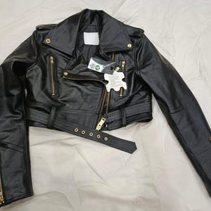 May include: Black leather jacket with a classic biker style. Features a notched collar, asymmetrical zip closure, and multiple zippered pockets. Includes a belt at the waist and gold-tone hardware. The jacket has long sleeves with zippered cuffs.