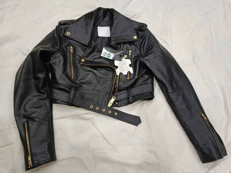 May include: Black leather jacket with a classic biker style. Features a notched collar, asymmetrical zip closure, and multiple zippered pockets. Includes a belt at the waist and gold-tone hardware. The jacket has long sleeves with zippered cuffs.