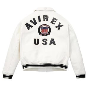 K&ouml;nnte beinhalten: Wei&szlig;e Lederjacke mit schwarz-wei&szlig; gestreiften B&uuml;ndchen und Saum. Auf dem R&uuml;cken steht das Wort "AVIREX" &uuml;ber einem kreisf&ouml;rmigen Emblem mit der amerikanischen Flagge und dem Wort "USA" darunter.