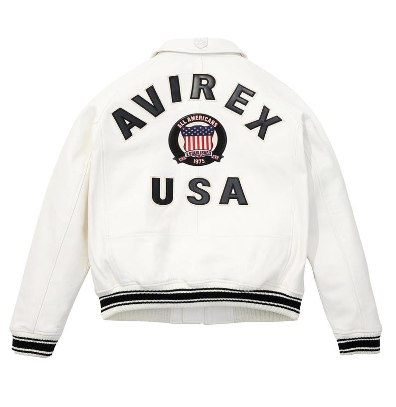 K&ouml;nnte beinhalten: Wei&szlig;e Lederjacke mit schwarz-wei&szlig; gestreiften B&uuml;ndchen und Saum. Auf dem R&uuml;cken steht das Wort "AVIREX" &uuml;ber einem kreisf&ouml;rmigen Emblem mit der amerikanischen Flagge und dem Wort "USA" darunter.