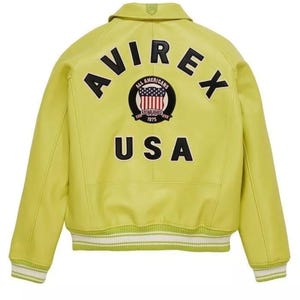 K&ouml;nnte beinhalten: Eine chartreusefarbene Jacke mit dem Wort "AVIREX" in schwarzen Buchstaben auf dem oberen R&uuml;cken. Darunter befindet sich ein kreisf&ouml;rmiges Emblem mit "ALL AMERICANS" und "ESTABLISHED 1975" und dem Wort "USA". Die Jacke hat einen wei&szlig;-gr&uuml;nen Streifen.