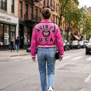 May include: A person wearing a pink leather jacket with the word "AVIREX" and "USA" on the back, along with a circular emblem. The jacket has white and black accents. The person is wearing blue jeans and white sneakers, walking on a city street.