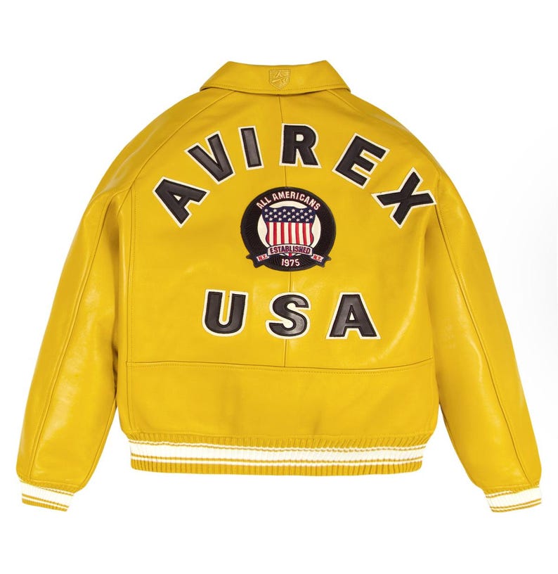 K&ouml;nnte beinhalten: Eine gelbe Lederjacke mit dem Wort "AVIREX" in schwarzen Buchstaben auf dem oberen R&uuml;cken. Darunter befindet sich ein kreisf&ouml;rmiger Aufn&auml;her mit der Aufschrift "ALL AMERICANS ESTABLISHED 1975" und einem US-Flaggen-Design. Das Wort "USA" steht unter dem Aufn&auml;her.
