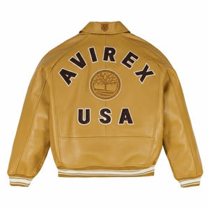 K&ouml;nnte beinhalten: Senfgelbe Lederjacke mit dem Wort "AVIREX" in braunen Buchstaben auf dem R&uuml;cken, &uuml;ber einem kreisf&ouml;rmigen Emblem und dem Wort "USA". Die Jacke hat einen gerippten Saum und B&uuml;ndchen mit wei&szlig;en und beigefarbenen Streifen.