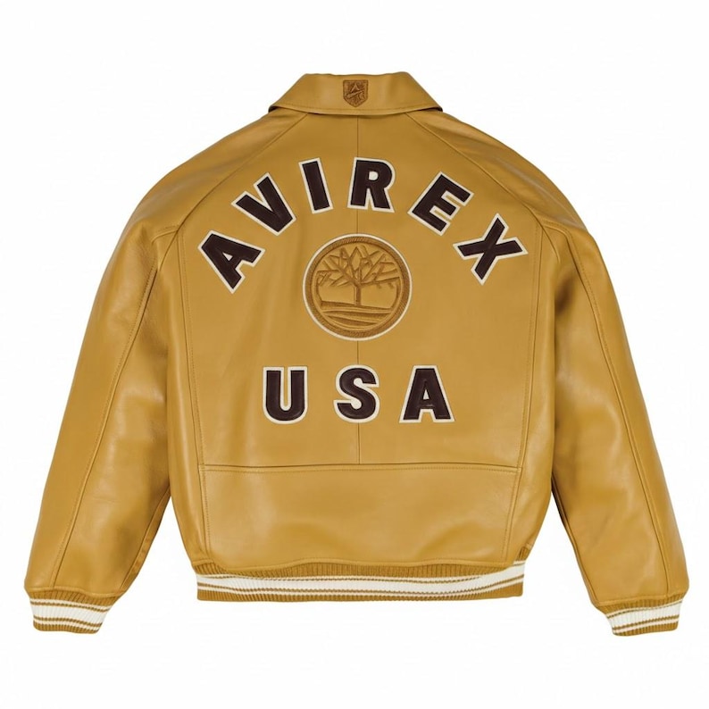 K&ouml;nnte beinhalten: Senfgelbe Lederjacke mit dem Wort "AVIREX" in braunen Buchstaben auf dem R&uuml;cken, &uuml;ber einem kreisf&ouml;rmigen Emblem und dem Wort "USA". Die Jacke hat einen gerippten Saum und B&uuml;ndchen mit wei&szlig;en und beigefarbenen Streifen.