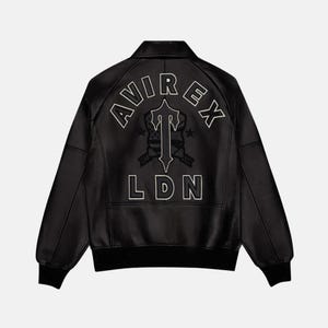 K&ouml;nnte beinhalten: Schwarze Lederjacke mit dem wei&szlig;en Schriftzug "AVIREX LDN" auf dem R&uuml;cken. Die Jacke hat einen klassischen Bomberstil mit geripptem Kragen und B&uuml;ndchen. Das Design beinhaltet ein stilisiertes Emblem.