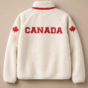 May include: Cream-coloured fleece jacket with a red collar and the word "CANADA" in red block letters across the back. Red maple leaf emblems are on each shoulder. A warm, comfortable outerwear garment.