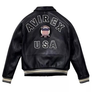K&ouml;nnte beinhalten: Schwarze Lederjacke mit dem Wort "AVIREX" in wei&szlig;en Buchstaben &uuml;ber den Schultern und "USA" darunter. Ein kreisf&ouml;rmiger Aufn&auml;her mit der amerikanischen Flagge befindet sich zwischen den W&ouml;rtern. Die Jacke hat einen gerippten Kragen und B&uuml;ndchen.