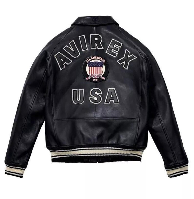 K&ouml;nnte beinhalten: Schwarze Lederjacke mit dem Wort "AVIREX" in wei&szlig;en Buchstaben &uuml;ber den Schultern und "USA" darunter. Ein kreisf&ouml;rmiger Aufn&auml;her mit der amerikanischen Flagge befindet sich zwischen den W&ouml;rtern. Die Jacke hat einen gerippten Kragen und B&uuml;ndchen.