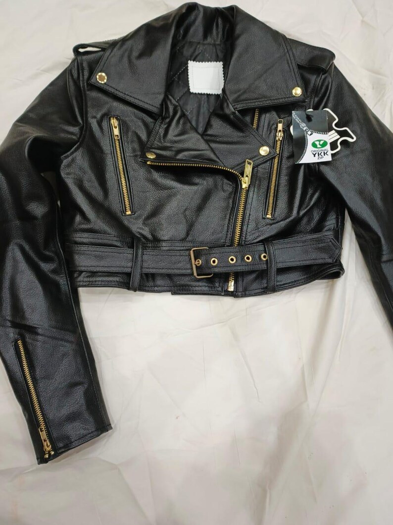 May include: Black leather jacket with a notched collar, asymmetrical zipper closure, and a belted waist. The jacket features gold-tone hardware, including zippers and snaps. A small tag with the text "YKK" is attached.