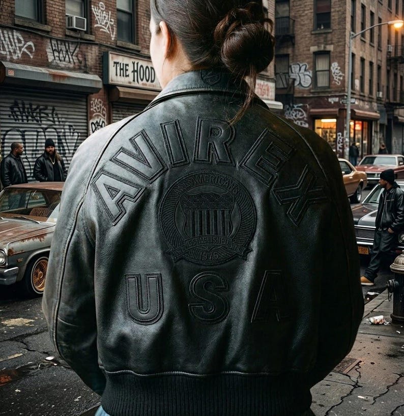 May include: Black leather bomber jacket with the word "AVIREX" across the back. The jacket features a circular emblem with the words "AMERICAN" and "ESTABLISHED 1975" and the letters "USA" below the emblem. The jacket is worn in an urban setting.