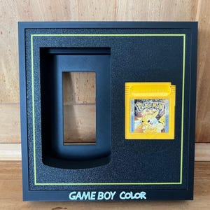 Peut inclure: Un cadre de présentation rectangulaire noir avec une cartouche de jeu vidéo Pokemon jaune sur le côté droit et une découpe rectangulaire à gauche. Le cadre a une fine bordure jaune et le texte GAME BOY COLOR imprimé en blanc en bas.