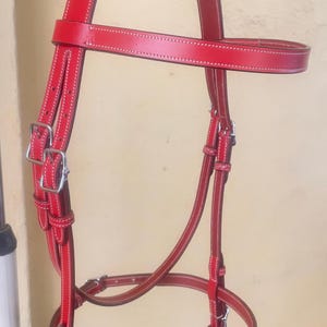 Puede incluir: Una brida de caballo de cuero rojo brillante con costuras blancas en contraste en la cabezada, la frontalera y la muserola. La brida incluye hebillas y herrajes de metal plateado para el ajuste, con múltiples correas y trabillas.