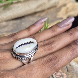May include: A silver ring featuring a polished white cowrie shell set in a metal bezel. The ring band has a decorative beaded edge detail surrounding the oval shell, which displays a natural dark brown slit running through its centre.