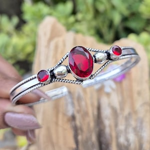 May include: A silver cuff bracelet featuring a large oval faceted red gemstone in the centre, flanked by two smaller round faceted red gemstones and two smooth silver spheres, all set within a twisted rope-textured silver metal band.