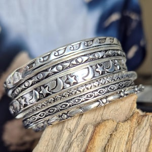 Op de afbeelding: Een zilverkleurige ring met meerdere banden bestaande uit vijf gestapelde ringen. De banden vertonen diverse reliëfpatronen, waaronder herhalende maansikkels, sterren, geometrische vormen en getextureerde lijnen, wat zorgt voor een gelaagd uiterlijk.