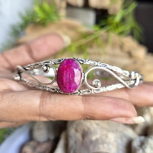 May include: A silver cuff bracelet featuring an oval-shaped, faceted magenta-pink gemstone at the center. The metal band displays an intricate, textured pattern with decorative swirling wire elements on both sides of the central stone setting.