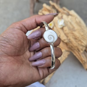 May include: A silver cuff bracelet featuring a circular white shiva eye shell inlay. The shell displays a natural spiral pattern in shades of brown and white. The metal band is smooth, polished, and open-ended, showcasing a clean, metallic finish.