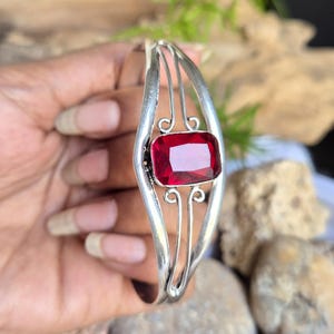 May include: A silver cuff bracelet featuring a central rectangular faceted red gemstone. The metal band has a split design with curved wire accents above and below the stone, creating an open frame structure around the vibrant red centre piece.