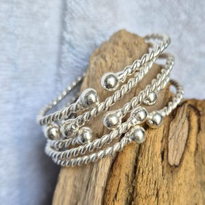 Puede incluir: Una pulsera de brazalete plateado con un diseño de cuerda trenzada de múltiples hilos. La pulsera está construida con varias bobinas metálicas interconectadas, cada una terminando en una pequeña cuenta esférica lisa y pulida en los extremos.