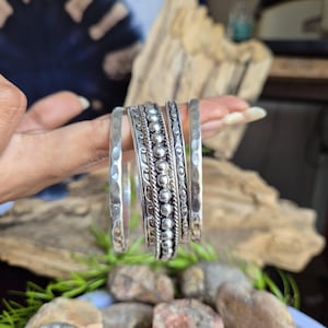 May include: A set of four silver-toned metal bangles. Two bangles feature a hammered texture, one displays a central row of raised spheres flanked by twisted rope detailing, and one has an intricate engraved pattern with small oval accents.