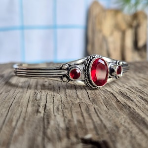 Puede incluir: Un brazalete de tono plateado con una piedra preciosa roja central de corte ovalado rodeada por un borde de metal trenzado, flanqueada por dos piedras preciosas rojas redondas más pequeñas a cada lado, sobre una banda de metal pulido.
