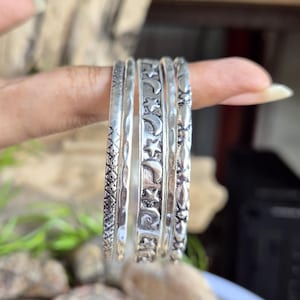 May include: A set of five silver-toned metal bangles featuring distinct textures and patterns. One band displays a repeating crescent moon and star motif, while others feature geometric cross-hatching, smooth hammered surfaces, and floral-like embossed details.