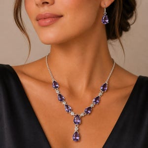 Puede incluir: Un conjunto de collar y pendientes de plata con piedras preciosas moradas en forma de pera. El collar tiene un diseño central de gota con pequeños detalles florales entre cada piedra, y el pendiente visible presenta una piedra morada a juego.