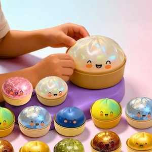May include: A collection of colorful, smiling dumpling-shaped toys. Each toy has a different color and glittery finish, with a happy face design. The toys are in a yellow container, and a child's hand is holding one of the larger toys.